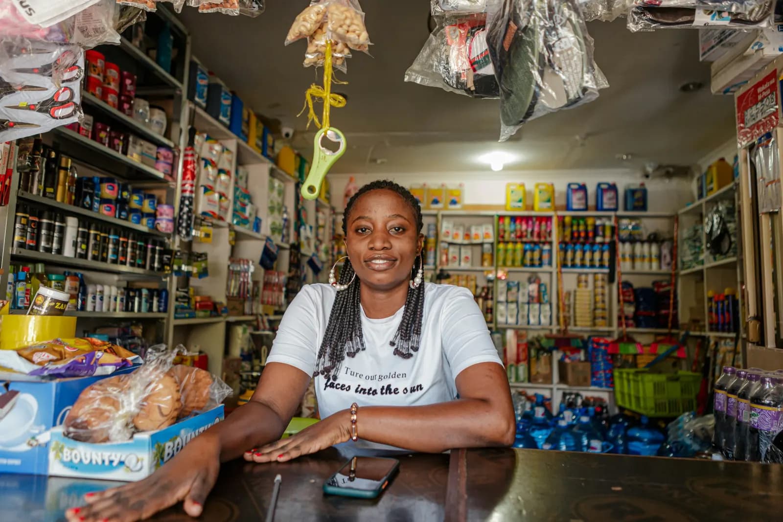 A shopkeeper smiling behind the counter of her general store in Dar es Salaam, a phone on the counter beside her.
