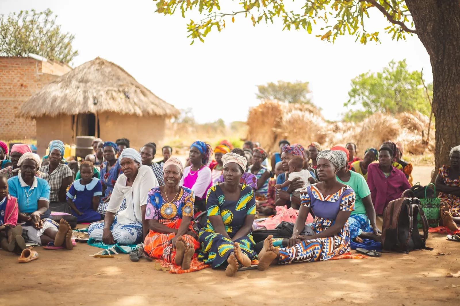 Women seated together on the ground in a village, thatched huts behind them.