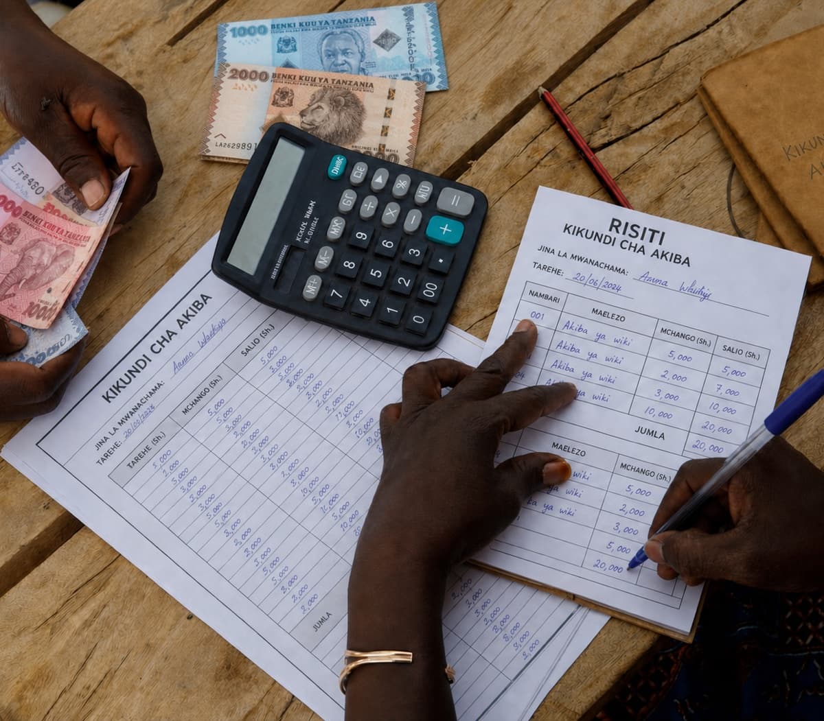 Overhead view of a collection on a wooden table: one pair of hands counts Tanzanian shilling banknotes while another writes in a handwritten RISITI receipt, beside a desk calculator and a handwritten ledger ruled into date, amount and balance columns.